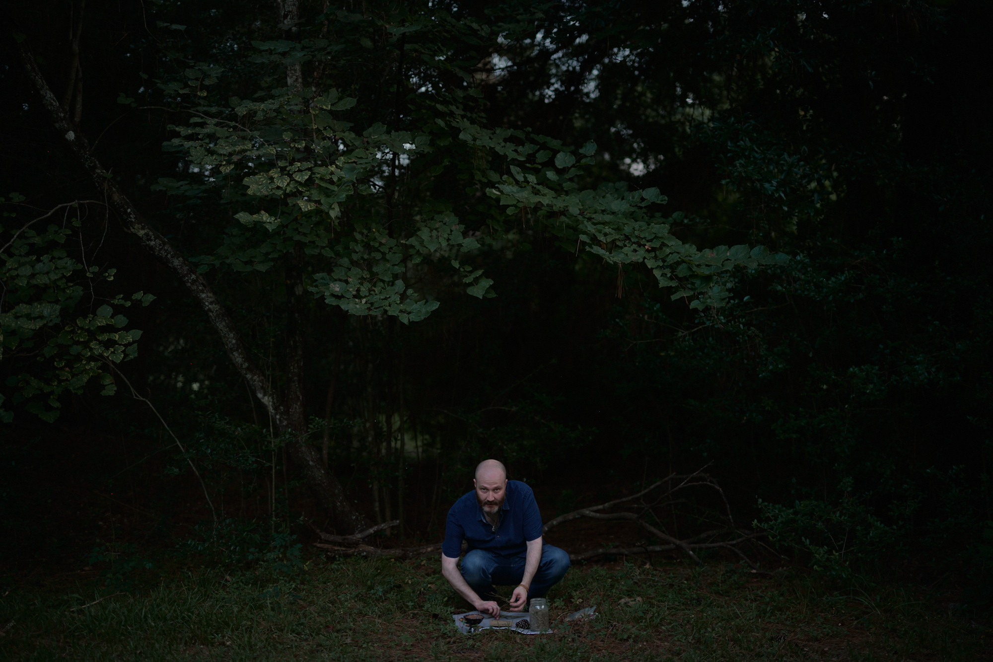 Photo of a man kneeling in the forest with a cloth holding tools and plants on the ground in front of him