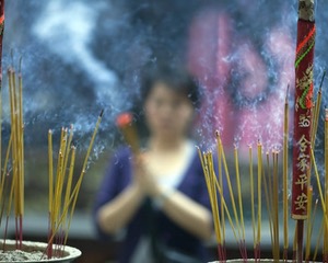 Photo of stick incense burning in the foreground, with a woman holding more incense in the blurred background